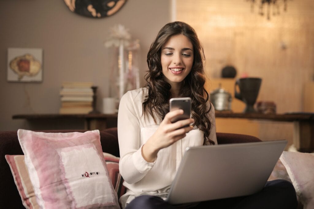 Woman Online Shopping While Looking at Phone With Laptop on Lap